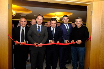 First image shows a large group of about 18 professionals in business attire standing together in front of a banner reading Antalya 2025 OKSİAD 2. Genel İstişare Toplantısı with a circular logo, in an indoor venue with wooden flooring and potted plants. Second image depicts a man in a checkered suit and striped tie speaking into a microphone on a podium against a beige wall. Third image features two men in suits holding a framed plaque with a red circular emblem, standing in front of a white backdrop. Fourth image captures five men in formal wear cutting a red ribbon at an entrance to a warmly lit room with ornate ceiling and chandelier.