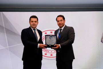 First image shows a large group of about 18 professionals in business attire standing together in front of a banner reading Antalya 2025 OKSİAD 2. Genel İstişare Toplantısı with a circular logo, in an indoor venue with wooden flooring and potted plants. Second image depicts a man in a checkered suit and striped tie speaking into a microphone on a podium against a beige wall. Third image features two men in suits holding a framed plaque with a red circular emblem, standing in front of a white backdrop. Fourth image captures five men in formal wear cutting a red ribbon at an entrance to a warmly lit room with ornate ceiling and chandelier.