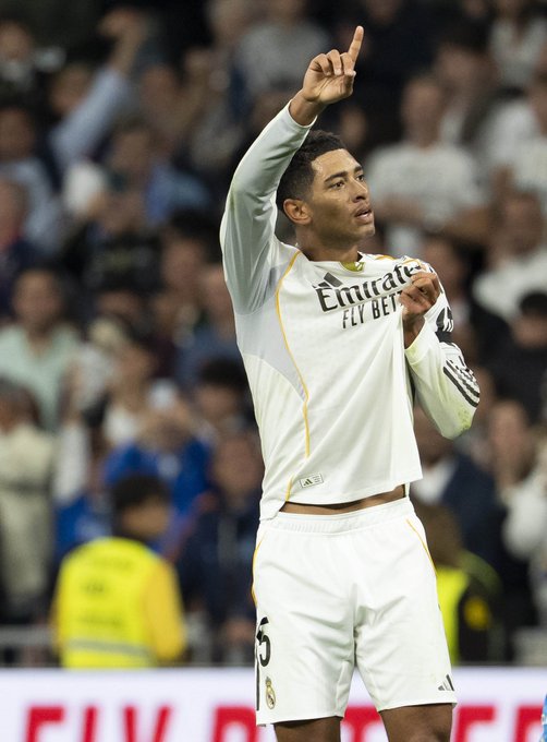 A male soccer player with short dark hair stands on a stadium field during a match, raising his right arm in celebration with index finger pointed up, wearing a white long-sleeve Real Madrid jersey with Emirates sponsor logo, white shorts numbered 15, and white socks, surrounded by blurred stadium seating and other players in similar uniforms.