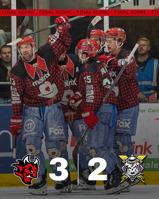 Group of male ice hockey players in red plaid jerseys with sponsor logos like Main and Matrix and numbers such as 15 wearing red helmets and blue jeans over pads raising hockey sticks in celebration on ice rink with scoreboard showing final score Devils 3 Storm 2 team logos of devil and dog mascot visible in background fans and officials in stands.