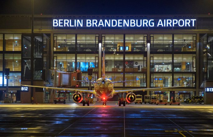 A commercial airplane positioned on the tarmac at night facing the modern terminal building with the illuminated sign BERLIN BRANDENBURG AIRPORT above large glass windows and multiple gates visible in the background including ground equipment and runway markings nearby.