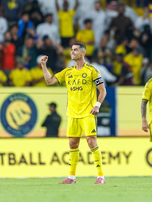 Cristiano Ronaldo stands on a soccer field wearing a yellow Al-Nassr jersey with sponsor logos, black armband, and red cleats, raising his right fist in celebration while smiling, surrounded by blurred stadium crowd in yellow attire and advertising boards.
