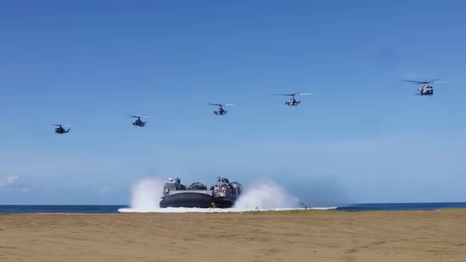 Multiple military helicopters in formation fly over a large amphibious landing craft creating a water spray as it approaches a sandy beach shore in a coastal area with clear blue skies.