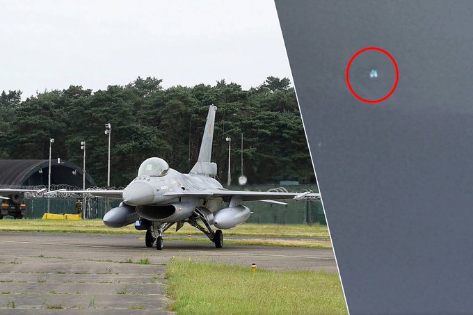 Gray fighter jet with cockpit canopy and undercarriage visible, parked on a grassy runway area near yellow barriers and fencing. Surrounding area includes green trees, gray buildings, and light poles under overcast sky. Red circle highlights a small indistinct object in the upper sky portion.