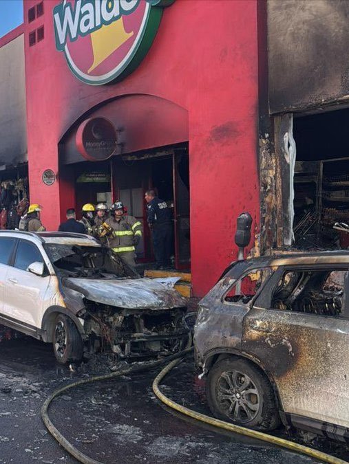 First image shows a red Waldos store building with visible fire damage and smoke emerging from broken windows and doors while several firefighters in yellow gear and helmets stand near the entrance connected to hoses. A white SUV and a black SUV both heavily damaged with fire and debris are crashed into the storefront area with hoses laid on the ground. Second image depicts the same Waldos store from a side angle with more smoke billowing out and structural collapse visible on the facade as a police officer in uniform stands beside the burned white SUV and scattered wreckage including metal debris and a red fire extinguisher nearby.