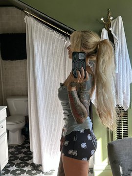 Woman with long blonde hair in pigtails takes mirror selfie in bathroom, wearing gray tank top and black shorts with elephant prints, tattoos on arms and legs visible, standing near white shower curtain, green walls, toilet, and tiled floor. Same woman leans against beige Jeep Wrangler SUV on dirt road in wooded area, dressed in black top, pants, boots, and cap, with off-road tires, power lines, and trees in background.