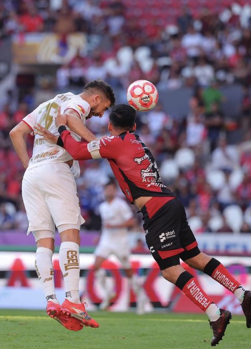 Two soccer players in uniforms compete mid-air for a red and white ball during a match on a green field with stadium seating and spectators in the background, one player in white jersey number 10 reaching with arm extended while the other in red and black kit jumps from behind.