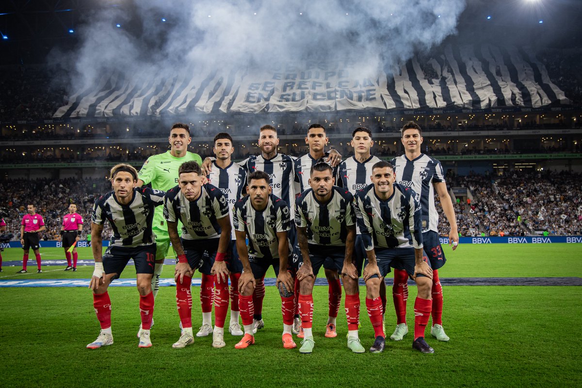 Eleven male soccer players in black and white striped jerseys with blue details, shorts, red socks, and cleats pose on a green field, some kneeling with hands on knees, others standing behind, in a large stadium filled with seats and a smoky atmosphere, a massive vertical banner of team stripes and text visible in the background, referees in yellow and black uniforms stand nearby.