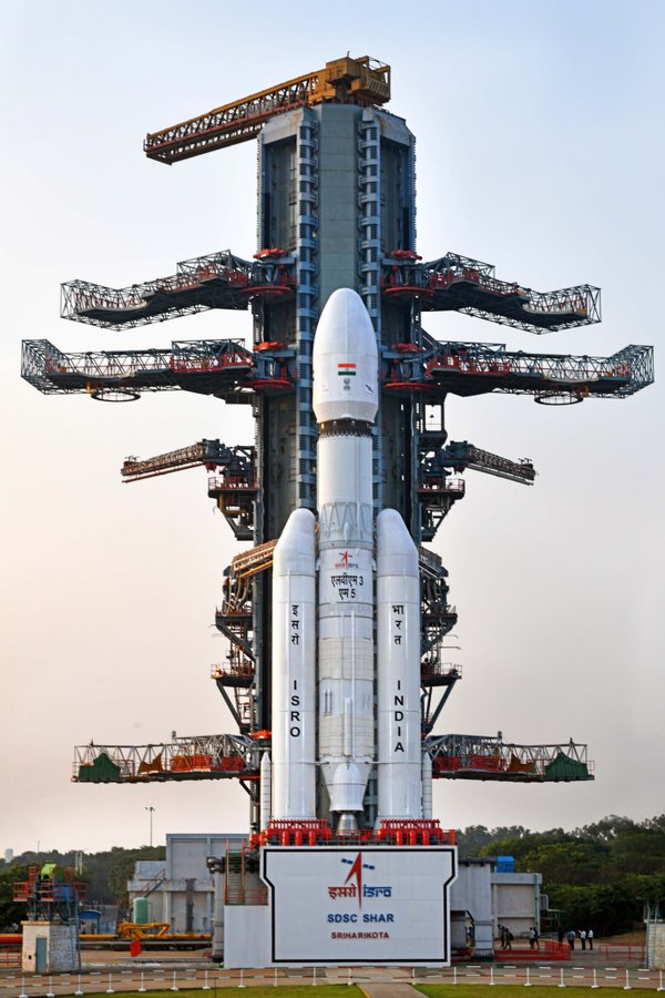 White rocket with Indian flag markings stands vertically on red launch platform surrounded by multiple levels of gray metal scaffolding and support structures under blue sky with green vegetation in background and ISRO sign at base