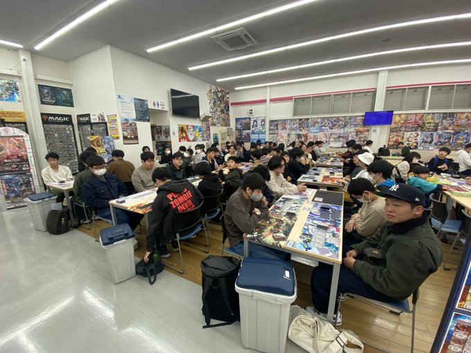 Indoor store room with white walls, large windows, air conditioning unit, and fluorescent lighting. Multiple rectangular tables arranged in rows, covered with green mats. Groups of men in casual clothing including hoodies, hats, and masks seated around tables, engaged in card games with decks, cards, and playmats visible. Shelves and displays on walls featuring trading card game posters, boxes, and merchandise. Trash bins and bags near tables. Blue stools and chairs around tables.