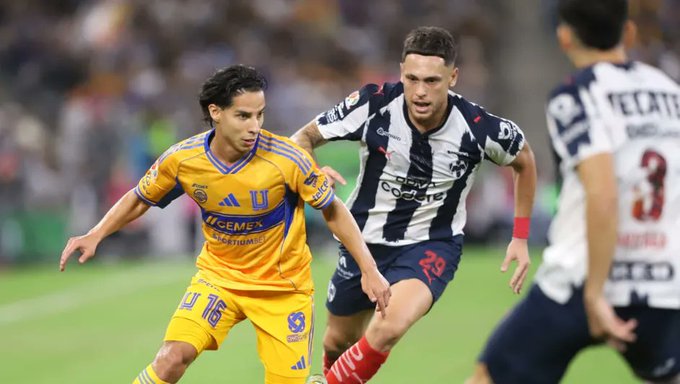 Two soccer players in action on a green field during a match. One player wears a yellow Tigres uniform with number 16 and blue accents, running with the ball. Another player in a white and blue striped Rayados uniform with number 23 pursues from behind. A referee in yellow and black is visible nearby. The stadium crowd appears in the blurred background.