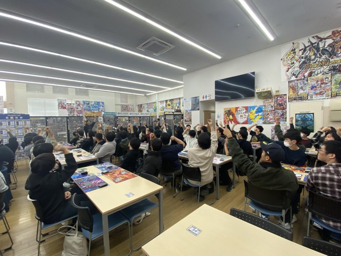 Indoor room with white walls and fluorescent lighting features trading card game shelves filled with boxed sets and posters of anime-style characters on walls. Large group of about 20-30 people mostly men in casual winter clothing like jackets and hats sit at rectangular white tables arranged in rows facing a front area. Participants hold up hands in rock-paper-scissors gestures during tournament. Air conditioning unit visible on wall and large flat-screen TV mounted above shelves. Floor is light wooden and chairs are simple black.