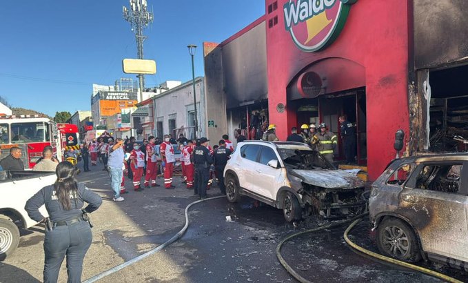 Scene outside a red Waldo’s store with a large green logo on a building facade showing fire damage and smoke. Fire trucks and emergency vehicles parked on the street with firefighters in red uniforms and helmets working near the entrance. A white SUV appears crashed and burned nearby. Police officer in gray uniform stands observing. Street with poles and signs in background under clear sky.