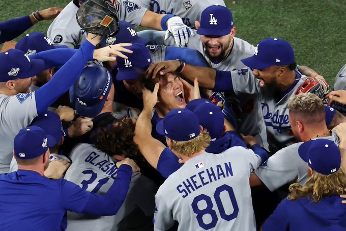 Group of Los Angeles Dodgers baseball players in blue jerseys and caps piling on top of each other on green field grass in celebration, with visible names like Sheehan 80 and Glasnow 31 on backs, Yoshinobu Yamamoto at center being hugged, all smiling and wearing gloves and helmets.