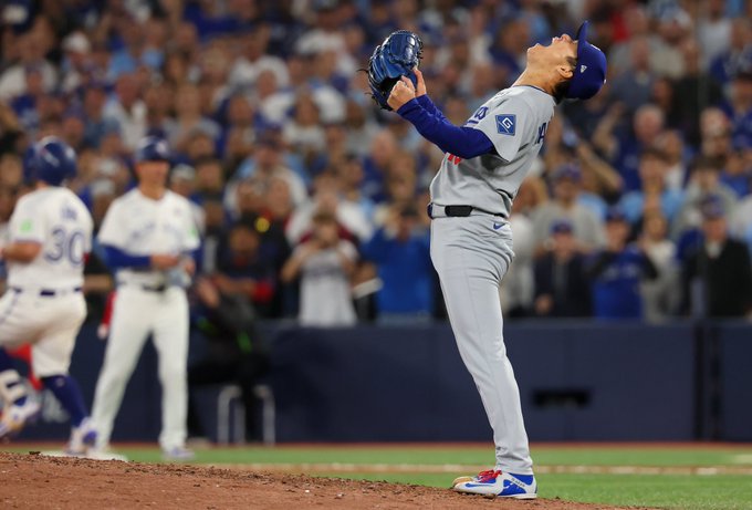 Jugador de beisbol con uniforme azul y gris de los Dodgers de Los Angeles celebra levantando una pelota en el guante derecho en el monticulo durante un juego de la Serie Mundial frente a una multitud en el estadio con otros jugadores y arbitros visibles en el campo y gradas llenas.