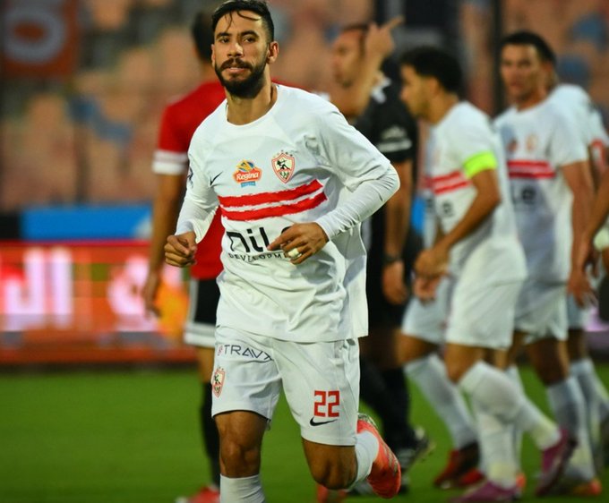 Soccer player Nasser Maher in white long-sleeve jersey with red stripes and sponsor logos including Zamalek emblem runs on green field during match wearing number 22 shorts and red cleats surrounded by teammates in matching uniforms some in red shirts.