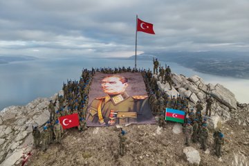 First image shows a large portrait of Mustafa Kemal Ataturk in military uniform placed on rocky mountain terrain near a lake with Turkish and Azerbaijani flags waving soldiers in camouflage uniforms standing in formation around it under cloudy sky. Second image depicts soldiers in camouflage on a rocky mountain peak overlooking a lake with Turkish and Azerbaijani flags planted nearby in a group formation. Third image features soldiers in camouflage uniforms positioned among rocks and sparse vegetation on a mountainous area with shadows cast on the ground under partly cloudy sky. Fourth image illustrates a group of soldiers in camouflage helmets and uniforms standing in a circle around a rectangular stone-lined area with a central object possibly a memorial or marker holding rifles and one soldier pointing with a stick.
