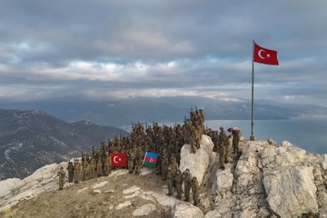 First image shows a large portrait of Mustafa Kemal Ataturk in military uniform placed on rocky mountain terrain near a lake with Turkish and Azerbaijani flags waving soldiers in camouflage uniforms standing in formation around it under cloudy sky. Second image depicts soldiers in camouflage on a rocky mountain peak overlooking a lake with Turkish and Azerbaijani flags planted nearby in a group formation. Third image features soldiers in camouflage uniforms positioned among rocks and sparse vegetation on a mountainous area with shadows cast on the ground under partly cloudy sky. Fourth image illustrates a group of soldiers in camouflage helmets and uniforms standing in a circle around a rectangular stone-lined area with a central object possibly a memorial or marker holding rifles and one soldier pointing with a stick.