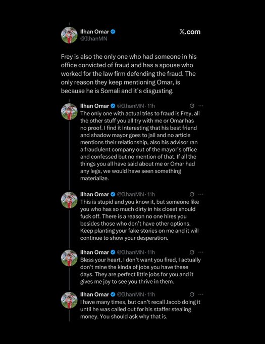 Ilhan Omar stands at a podium speaking with mouth open and right hand raised pointing upward wearing a black headscarf gray blazer black top gold bracelet and ring against a blue sky background with a microphone visible.