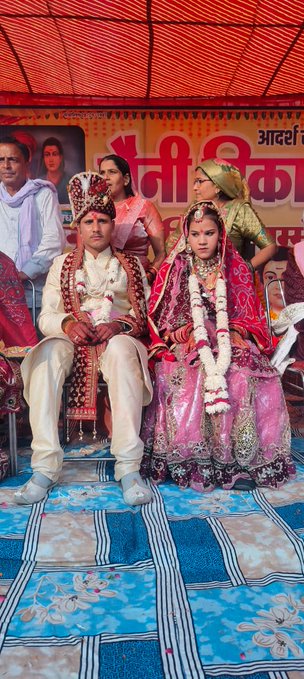 First image shows a large group of people seated on the floor under an orange tent with Hindi banners reading Saini Vikas Sangathan. Men and women in white and colorful traditional outfits including turbans and sarees sit in rows facing a stage with speakers and a table. Some wear garlands and jewelry. The setting includes patterned rugs and poles supporting the tent. Second image focuses on a bride and groom seated together; the bride in a pink sparkling lehenga with heavy necklace and the groom in cream sherwani with turban both holding hands and wearing garlands. Background has Hindi banner Anni Saini and portraits. Third image shows another couple; the bride in red saree with dupatta and the groom in white sherwani both with garlands seated under a blue and white banner mentioning Mini Sabha. Elderly attendees in traditional clothes surround them on chairs. Fourth image captures multiple couples in wedding attire seated in rows under a tent with yellow and purple banners for Shri Vikas Sangathan. Men in sherwanis and women in red and pink sarees with jewelry and garlands. A stage with microphone and table is visible along with patterned blue floor.