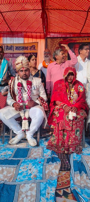 First image shows a large group of people seated on the floor under an orange tent with Hindi banners reading Saini Vikas Sangathan. Men and women in white and colorful traditional outfits including turbans and sarees sit in rows facing a stage with speakers and a table. Some wear garlands and jewelry. The setting includes patterned rugs and poles supporting the tent. Second image focuses on a bride and groom seated together; the bride in a pink sparkling lehenga with heavy necklace and the groom in cream sherwani with turban both holding hands and wearing garlands. Background has Hindi banner Anni Saini and portraits. Third image shows another couple; the bride in red saree with dupatta and the groom in white sherwani both with garlands seated under a blue and white banner mentioning Mini Sabha. Elderly attendees in traditional clothes surround them on chairs. Fourth image captures multiple couples in wedding attire seated in rows under a tent with yellow and purple banners for Shri Vikas Sangathan. Men in sherwanis and women in red and pink sarees with jewelry and garlands. A stage with microphone and table is visible along with patterned blue floor.