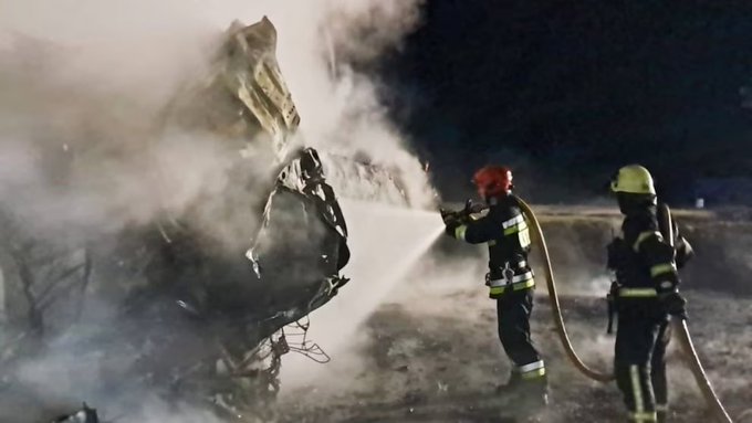 Two firefighters in full gear including helmets and yellow suits spray water from hoses onto a heavily damaged dark-colored vehicle with its front end crushed and deformed surrounded by thick white smoke billowing from the wreckage at night in an outdoor setting with some trees visible in the background.