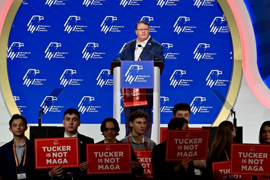 Tucker Carlson stands at a podium wearing a suit and tie speaking into a microphone with a blue circular backdrop featuring repeated eagle emblems. Several young men and women in formal attire sit and stand around holding red signs that read Tucker is not MAGA. Additional signs and American flags are visible in the audience area.