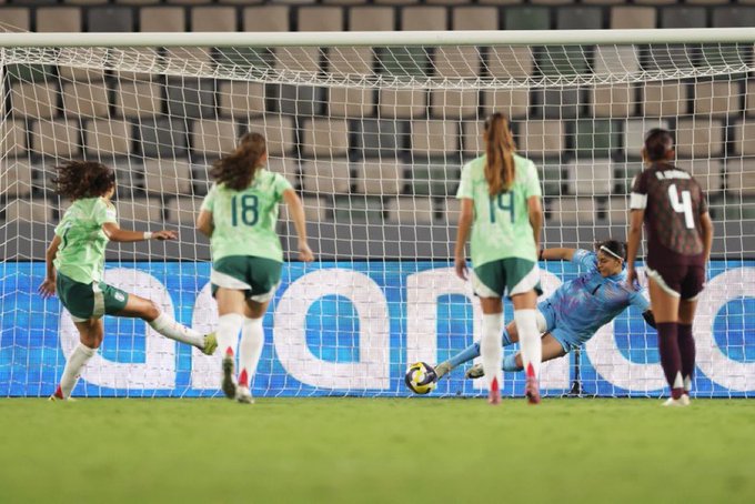 Four female soccer players in green uniforms numbered 18 and 19 attempt a goal kick towards a yellow soccer ball near the net while a goalkeeper in a maroon uniform numbered 4 dives to block it on a stadium field with blue sponsor banners.