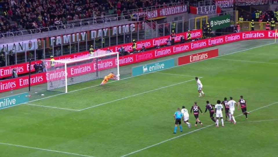 A soccer field scene shows goalkeeper Mike Maignan in a white and red kit diving low to his right with arms extended to save a shot, blocking the ball near the goal line marked with white paint. Several outfield players in black and red kits pursue the play around him, including one in green. The goal net bulges slightly behind, with a referee in yellow nearby. Stadium stands filled with spectators rise in the background, displaying banners for Family Day AC Milan and sponsor ads like Emirates and Etihad. Green grass covers the pitch under daylight.