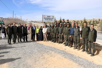 First image shows diverse group of people including women in headscarves and a young boy in white shirt planting and watering small pine saplings with green watering cans marked with forestry logo in dry soil near a wooden stake. Second image displays crowd gathered around white signboard reading Sehit Can Muharrem Ormani with Turkish flags and vehicles on dusty roadside in hilly terrain. Third image captures participants kneeling and digging to plant saplings in barren hillside landscape under blue sky with scattered bushes. Fourth image features line of officials in green uniforms and civilians standing formally beside plaque reading Sehit Can Muharrem Ormani against backdrop of arid hills and parked vehicles.