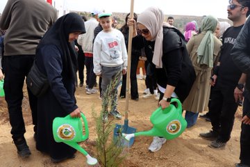 First image shows diverse group of people including women in headscarves and a young boy in white shirt planting and watering small pine saplings with green watering cans marked with forestry logo in dry soil near a wooden stake. Second image displays crowd gathered around white signboard reading Sehit Can Muharrem Ormani with Turkish flags and vehicles on dusty roadside in hilly terrain. Third image captures participants kneeling and digging to plant saplings in barren hillside landscape under blue sky with scattered bushes. Fourth image features line of officials in green uniforms and civilians standing formally beside plaque reading Sehit Can Muharrem Ormani against backdrop of arid hills and parked vehicles.