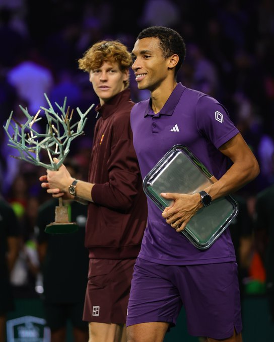 Two male tennis players stand side by side on an indoor court with a blurred crowd in the background. The player on the left, with curly red hair, wears a maroon jacket and pants, holding a trophy with branching metallic elements on a wooden base. The player on the right, with short dark hair, wears a purple polo shirt and shorts with Adidas and On logos, holding a silver rectangular plaque. Both smile and wear wristwatches.