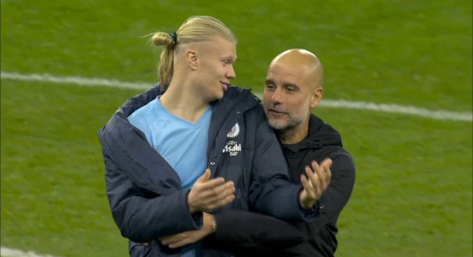 Erling Haaland with blonde ponytail in ponytail hairstyle wearing a dark jacket over a light blue shirt stands smiling on a green soccer field next to bald Pep Guardiola in black hoodie clapping hands with stadium white lines visible in background.