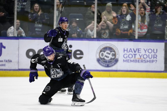 Two ice hockey players in purple Clan jerseys numbered 58 and another, wearing helmets and gloves, stand on the rink celebrating with sticks raised, one kneeling and the other standing, surrounded by blurred crowd spectators behind glass barriers, with team banners and sponsor ads like OSC on the boards.