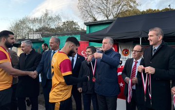 First image shows a man in a suit speaking into a microphone at a podium under a black canopy with Turkish flags banners and posters of Turkish figures and federation logos on a green background. Second image depicts two men one in a black coat and one in a blue jacket standing on a grassy soccer field with a ball at their feet surrounded by autumn trees and a crowd in the background. Third image captures a group of men in formal attire and sports jerseys shaking hands and holding medals under a canopy with Turkish flags and event banners. Fourth image features a line of men in suits cutting a red ribbon in front of a structure with Turkish flags on a paved area during sunset with trees nearby.