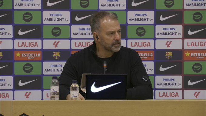 A man in a black jacket sits at a press conference table with a Nike laptop in front of him surrounded by sponsor banners including Spotify La Liga Estrella Damm Philips and Nike on a green backdrop with water bottles nearby.