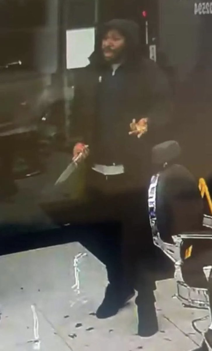 A man wearing a black hoodie and pants stands near a bus shelter at night holding a red cylindrical object in his right hand and possibly another item in his left near his face with a yellow chair and glass door visible behind him.