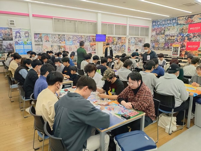 Numerous young men wearing casual clothes and masks sit at several rectangular tables arranged in rows inside a brightly lit indoor store space with wooden flooring and large windows. Colorful Pokemon cards and game mats cover the tables as participants lean forward engaged in gameplay. Walls are adorned with extensive posters and banners featuring Pokemon characters and artwork. A television screen is mounted on the wall displaying related content. The atmosphere appears focused and communal.