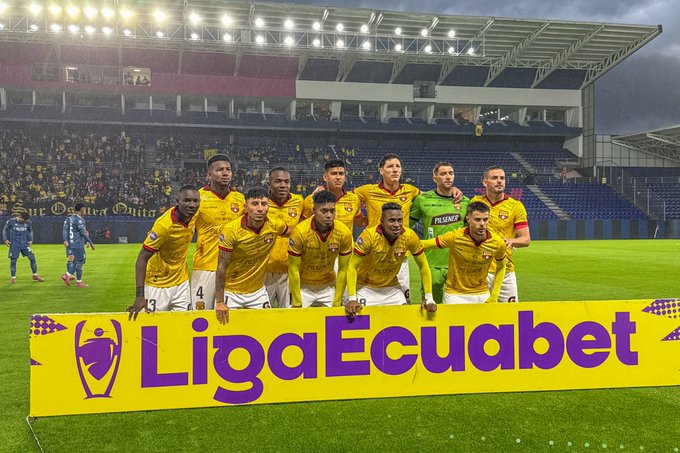 Group of eleven male soccer players in yellow and black striped uniforms with sponsor logos stand closely together on green grass field near sideline, arms around shoulders, smiling at camera. Goalkeeper in green jersey positioned among them. Purple banner with white text LigaPro and emblem lies on ground in front of team. Stadium seating with blue and purple sections and distant crowd visible in background under illuminated floodlights at dusk.