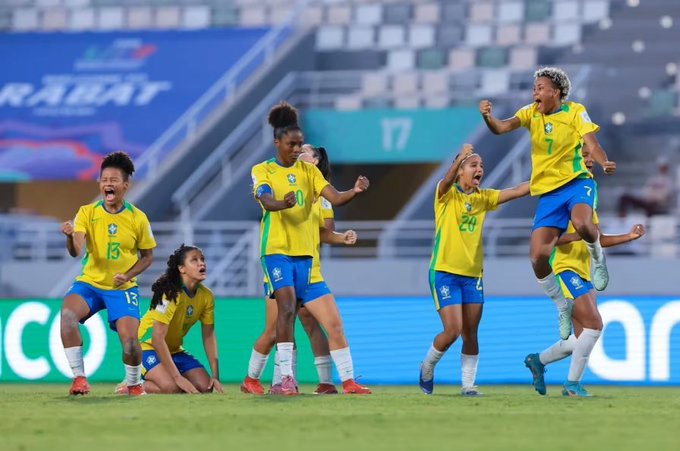 Group of young female soccer players in yellow jerseys and blue shorts celebrating a victory on a green soccer field. Several players jump with arms raised, one lifts another, showing joy with open mouths and smiles. Background includes blue stadium seating, a blue banner, and green turf with white lines. Numbers like 17 and 20 visible on jerseys.