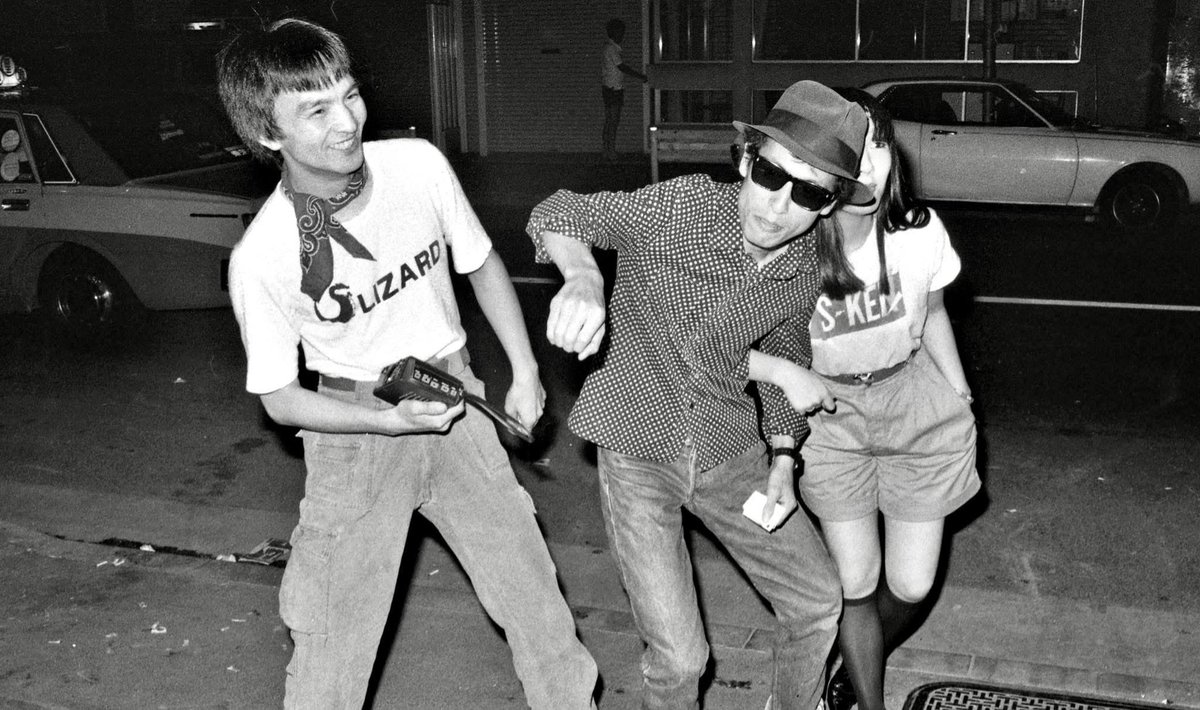 dudleytt's tweet photo. 旧友の作家・片山喜康と話してたら、“Tokyo Rockers”時代にLIZARDのベーシストだったワカ（若林一彦）さんの話になった。
ふたりとも浅草界隈の出身で、ワカさんの実家は観音様裏の千束通りにある花屋“花新”。片山氏いわく「ワカさんは寡黙だったけど、一本気でまさに江戸っ子だったよね」と。

今月は映画化で話題の『ストリート・キングダム』の原作者、地引雄一が送ってくれた当時の写真の中から、ワカさんの姿を探してみた。
探しながら“Tokyo Rockers”を思い返すと、FRICTION、Mr.KITE、MIRRORSのメンバーは高円寺を中心とした中央線沿線の住人が多かったが、LIZARDはモモヨが尾久、ワカさんが浅草、私が代々品川、ドラムのネズミ（富田英次）が十条と、東京を東西南北に散らばっていた。

この写真は1981年、新宿ロフト付近の路上で撮られたもので、ワカさんとs-kenTシャツを着た女の子と遊んでいるようなショット。44年前のことだから全く覚えはないけれど、なんとも楽しそう。
1981年といえば、デビューアルバム『魔都』がリリースされた年。前年に出たLIZARDのシングル「浅草六区（Asakusa Rock）」が好きだった。

〽仲見世を駆け抜けて　夢のカケラを探すのさ

この写真を見ていると、ちょうどその頃のワカさんは、この歌詞のように“夢のカケラ”を追いかけてニューウェイブの時代を駆け抜けていたのだと感じる。“和製ジャン＝ジャック・バーネル”なんて呼ばれていたのもその頃。

その後、LIZARDだけでなくさまざまなバンドをサポートしながら、やがてトラック運転手に転身。結婚して子どもも生まれ、平穏な生活を送っていた。
しかし東日本大震災後、反原発運動にのめり込み、2012年には原発再稼働に反対する市民団体の一員として、当時の野田佳彦首相官邸に乗り込んだり、デモの先頭に立ってパフォーマンスを行うなど、過激な行動をとるようになる。それは晩年まで続いたといからまさに一本気なワカさんらしい。

“Tokyo Rockers”時代に出会ったRockerたちの、その後の成り行きに思いを馳せるときが来たのかもしれない。
男から女へと性転換した者、新宗教の教祖のような存在になった者、何億も稼ぐ実業家となった者もいて、人生の行方はそれぞれに劇的で、驚きに満ちている。
だが半世紀近い時を経たいま、闘病中の仲間や、すでにこの世を去った者も少なくない。
年長者として生き延びた者としては、あの熱狂と混沌の時代を、彼ら彼女たちと共に生きられたこと——いま感謝しつつ胸の底に灯り続けている。