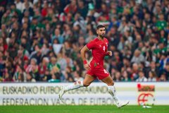 First image shows two male soccer players standing side by side on a green field in a stadium with a crowd in the background one in a green short-sleeved jersey with sponsor logos and the other in a red short-sleeved jersey numbered 4 both wearing athletic shorts white socks and cleats the green-jerseyed player has a wristband and the red one has a bag strap over shoulder. Second image depicts a male player in a red jersey numbered 4 running on the green field during a match with a blurred stadium crowd behind him wearing red shorts white socks with knee pads and white cleats legs in motion. Third image captures a male player in a red jersey numbered 4 walking and clapping on the green field with another player in green jersey clapping nearby both in athletic uniforms with stadium seating and field lines visible.