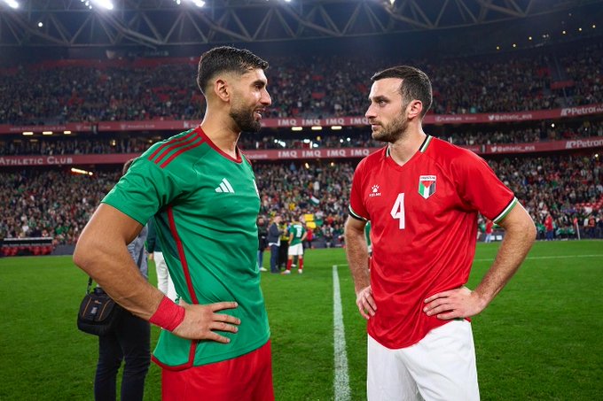 First image shows two male soccer players standing side by side on a green field in a stadium with a crowd in the background one in a green short-sleeved jersey with sponsor logos and the other in a red short-sleeved jersey numbered 4 both wearing athletic shorts white socks and cleats the green-jerseyed player has a wristband and the red one has a bag strap over shoulder. Second image depicts a male player in a red jersey numbered 4 running on the green field during a match with a blurred stadium crowd behind him wearing red shorts white socks with knee pads and white cleats legs in motion. Third image captures a male player in a red jersey numbered 4 walking and clapping on the green field with another player in green jersey clapping nearby both in athletic uniforms with stadium seating and field lines visible.