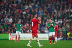 First image shows two male soccer players standing side by side on a green field in a stadium with a crowd in the background one in a green short-sleeved jersey with sponsor logos and the other in a red short-sleeved jersey numbered 4 both wearing athletic shorts white socks and cleats the green-jerseyed player has a wristband and the red one has a bag strap over shoulder. Second image depicts a male player in a red jersey numbered 4 running on the green field during a match with a blurred stadium crowd behind him wearing red shorts white socks with knee pads and white cleats legs in motion. Third image captures a male player in a red jersey numbered 4 walking and clapping on the green field with another player in green jersey clapping nearby both in athletic uniforms with stadium seating and field lines visible.