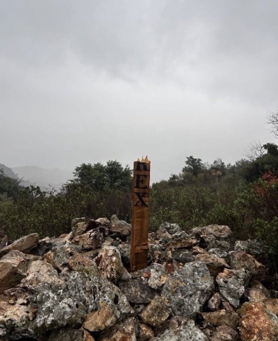 A wooden cross with the letters AVE GRATIA PLENA carved on it stands upright on a pile of rocks in a mountainous landscape with trees and clouds in the background. The cross is made of rough wooden beams and appears newly installed. Surrounding area includes bushes and distant views of valleys.