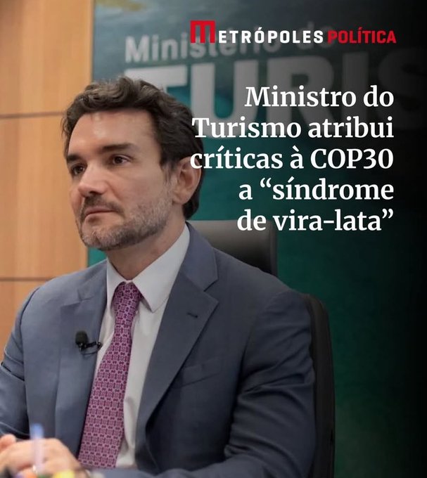 A man in a suit and tie sits at a desk in a formal setting with microphones, looking serious, behind him a backdrop with Metropoles Politica logo and text reading Ministro do Turismo atribui críticas à COP30 a síndrome de vira-lata.