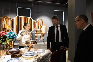 First image shows a group of eight formally dressed adults, including men in suits and women in traditional attire like hijab, standing in front of a large red banner promoting Ev Hali exhibition for family lifelong learning from May 3 to 18 2025, with logos of ministries and organizations, stone architecture in background. Second image displays various colorful traditional Turkish rugs and textiles hanging on gray walls in a gallery space, including patterned kilims, embroidered fabrics, and wooden furniture like stools and cushions arranged below. Third image captures three people interacting near exhibition displays: a woman in white holding a metal tray with pottery, a man in suit examining items, and another man in suit nearby, surrounded by hanging woven panels, chairs, flowers, and table settings with dishes. Fourth image features a man in dark suit standing alone in front of a red banner for Ev Hali exhibition detailing the event from May 3 to 18 2025 organized by MEB and related bodies, with descriptive text panels beside.