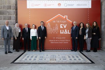 First image shows a group of eight formally dressed adults, including men in suits and women in traditional attire like hijab, standing in front of a large red banner promoting Ev Hali exhibition for family lifelong learning from May 3 to 18 2025, with logos of ministries and organizations, stone architecture in background. Second image displays various colorful traditional Turkish rugs and textiles hanging on gray walls in a gallery space, including patterned kilims, embroidered fabrics, and wooden furniture like stools and cushions arranged below. Third image captures three people interacting near exhibition displays: a woman in white holding a metal tray with pottery, a man in suit examining items, and another man in suit nearby, surrounded by hanging woven panels, chairs, flowers, and table settings with dishes. Fourth image features a man in dark suit standing alone in front of a red banner for Ev Hali exhibition detailing the event from May 3 to 18 2025 organized by MEB and related bodies, with descriptive text panels beside.