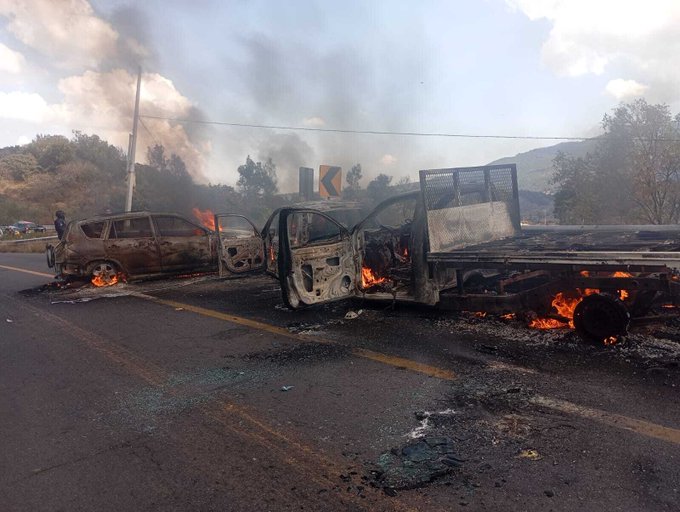 Multiple vehicles including a burned SUV and truck on a roadside with open doors flames and thick black smoke rising surrounded by utility poles trees and a mountainous background under a partly cloudy sky