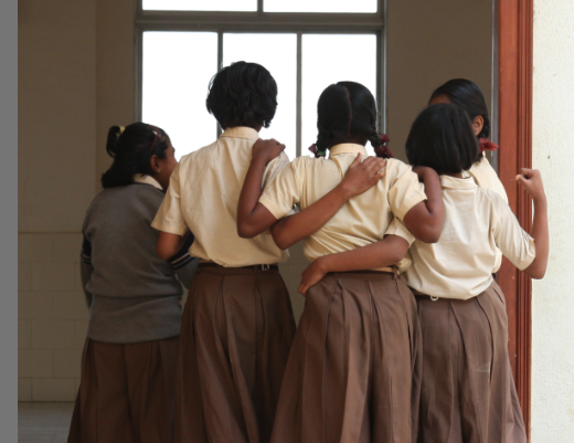Four young girls with dark hair in ponytails stand in a line facing away from the viewer inside a simple classroom with beige walls and a large window showing daylight outside. They wear matching school uniforms consisting of white shirts, gray sweaters, and brown skirts or trousers. The setting appears to be a school doorway with a wooden frame.